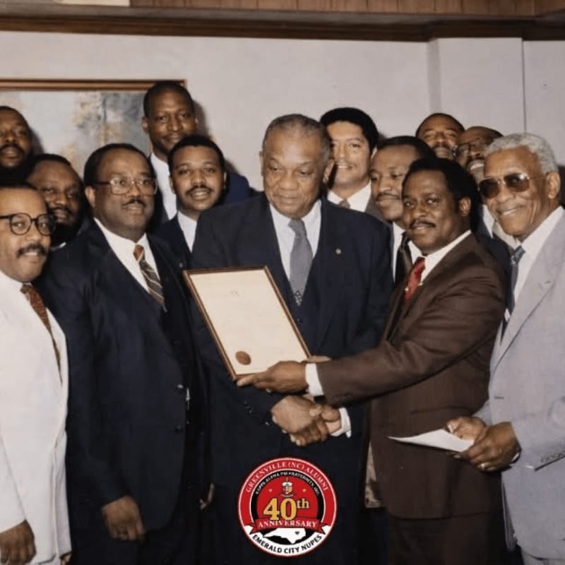 A group of men in suits gather around as one man presents a framed document, with a circular logo indicating a 40th anniversary in the foreground and a plain wall in the background.