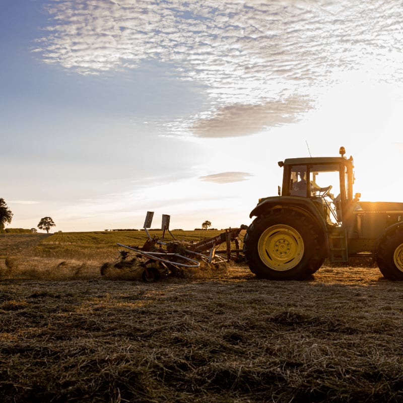 A tractor with a mower attachment works in a field of dry grass under a bright, cloudy sky.