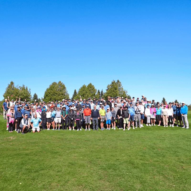 A large group of people stand on a grassy field with trees and a clear blue sky in the background.
