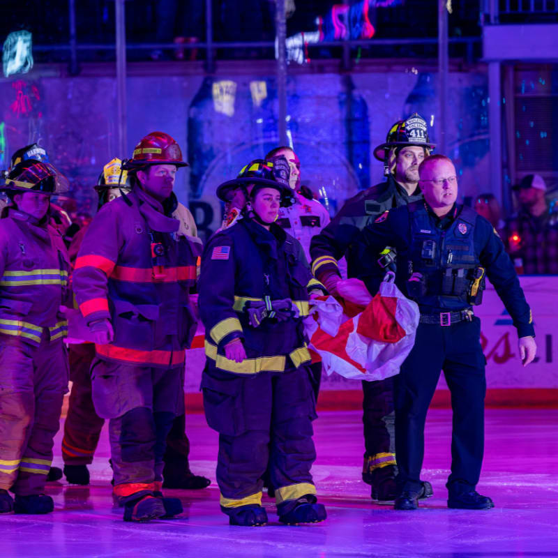 Firefighters in uniform stand on an ice rink in the foreground, while a blurred background shows lights and what appears to be a crowd.