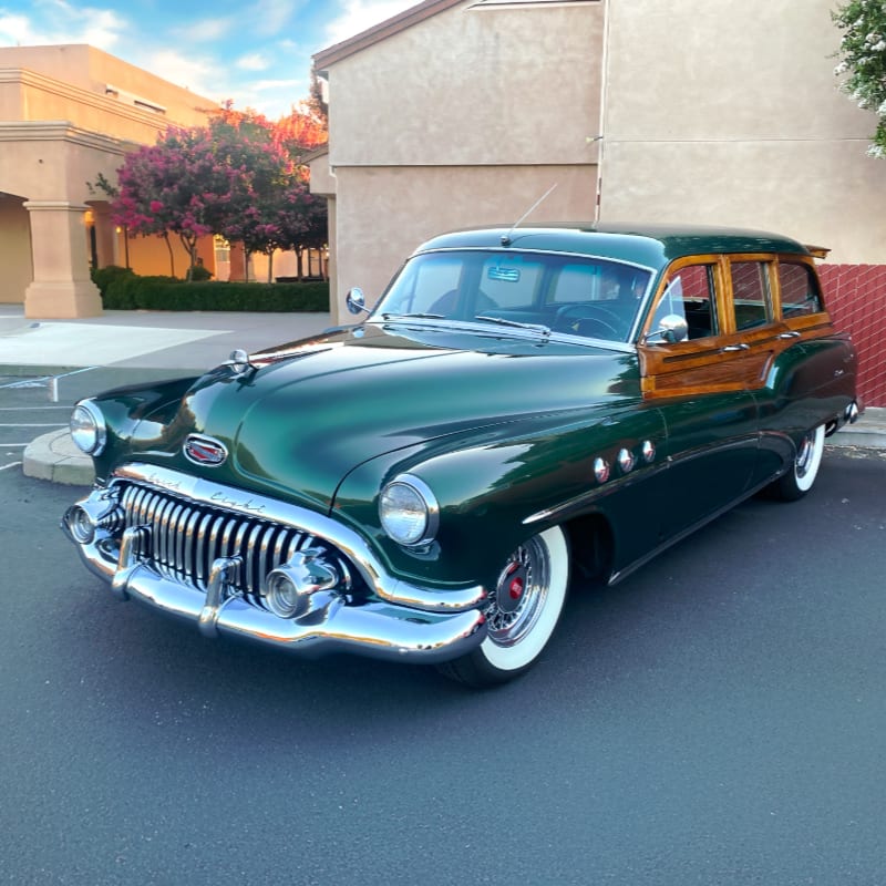 A gleaming green vintage station wagon with wooden paneling sits in a parking lot with a building and trees in the background.