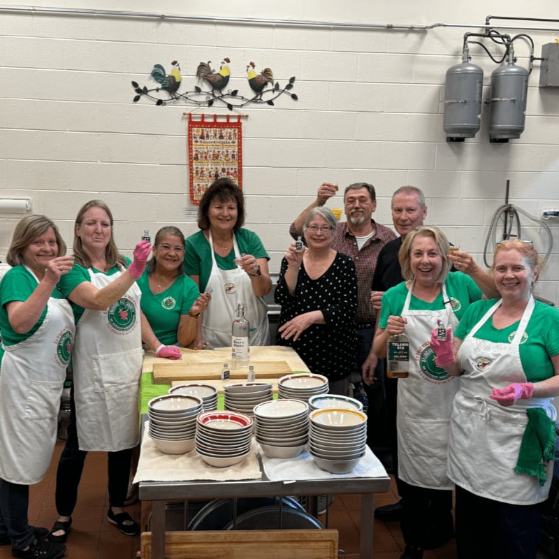 A group of people in green shirts and white aprons raise glasses in a kitchen with stacks of bowls on a table in the foreground and industrial equipment in the background.