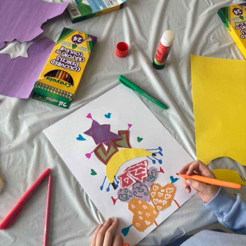 A child's hands are coloring a drawing on a white sheet of paper, surrounded by art supplies like colored pencils, glue sticks, and construction paper on a white tablecloth.