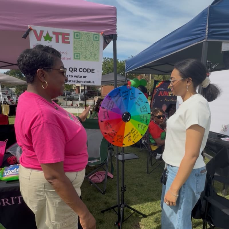 Two women stand facing each other in front of a colorful prize wheel, with tents and other people visible in the background.