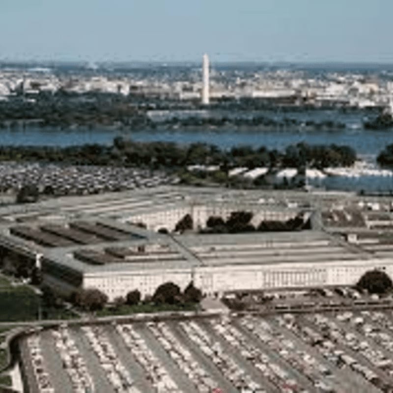 A vast parking lot surrounds the Pentagon in the foreground, with the Washington Monument and city skyline visible across the Potomac River in the background.