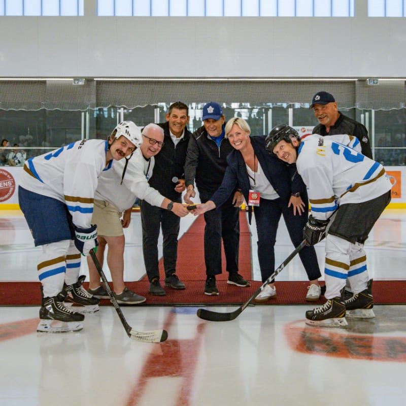 Seven people in hockey gear and casual attire gather on an ice rink with a red carpet and spectators in the background.
