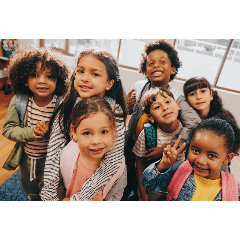 A diverse group of smiling children, some with backpacks, pose in front of a bright, blurred background.