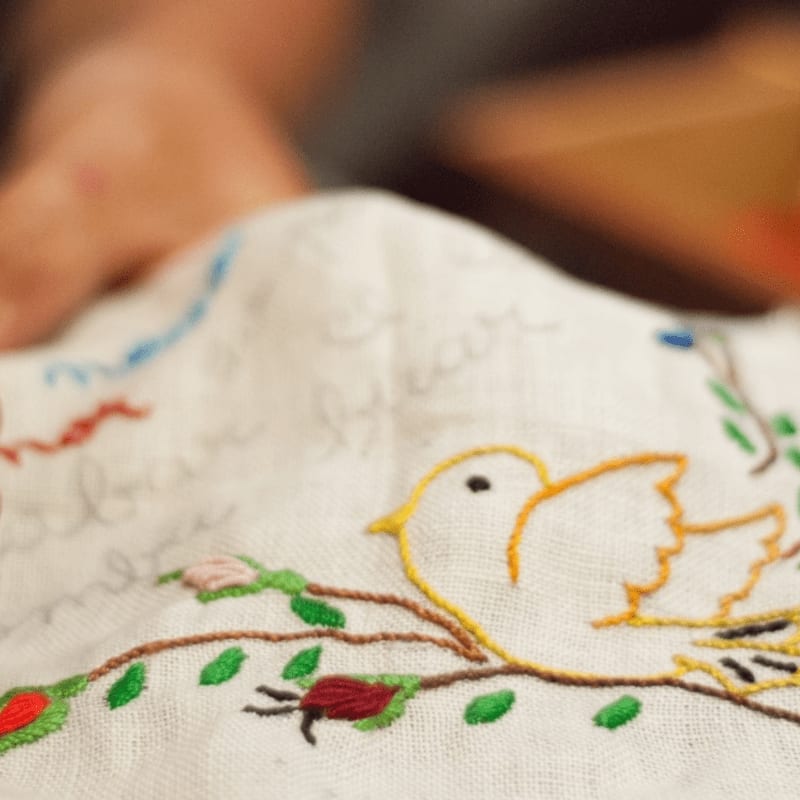 A close-up of a white embroidered fabric with a yellow bird on a branch in the foreground, and a blurred hand in the background.