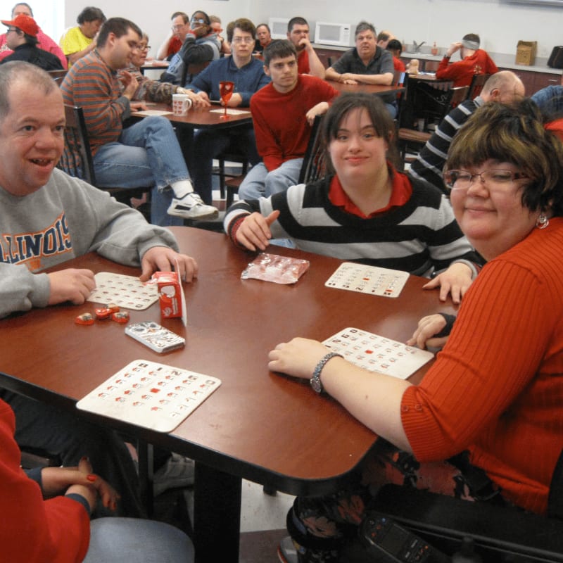 Four people with disabilities are playing bingo at a table in the foreground, while other people are seated at tables in the background.