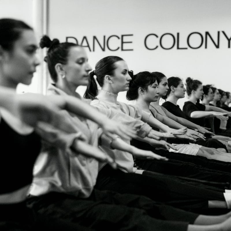 A line of women in a dance class with their arms extended forward, against a wall with "DANCE COLONY" written on it.