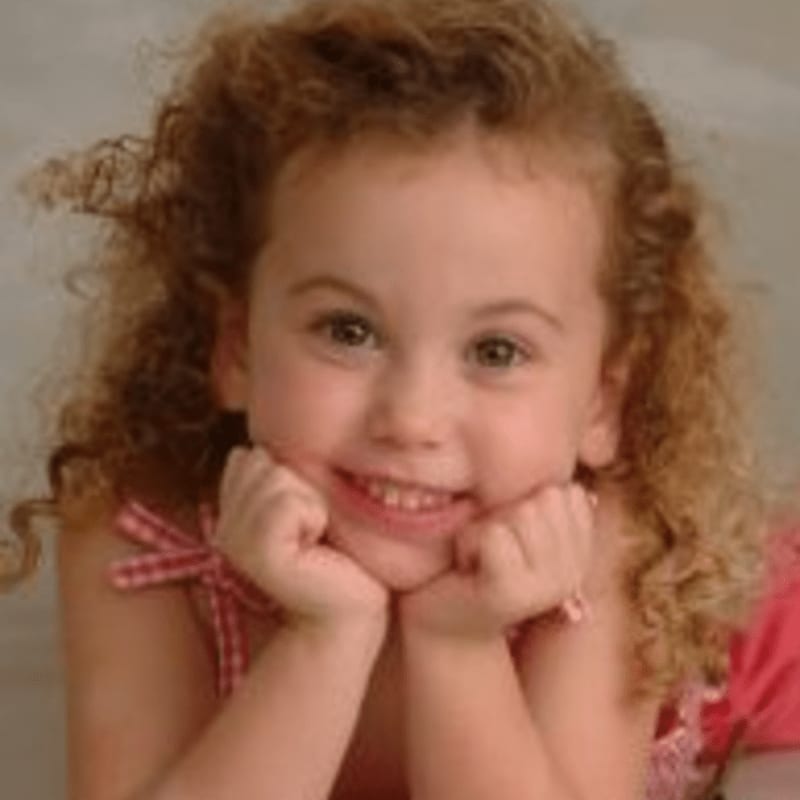 A young girl with curly hair rests her chin on her hands, smiling against a soft, blurred background.