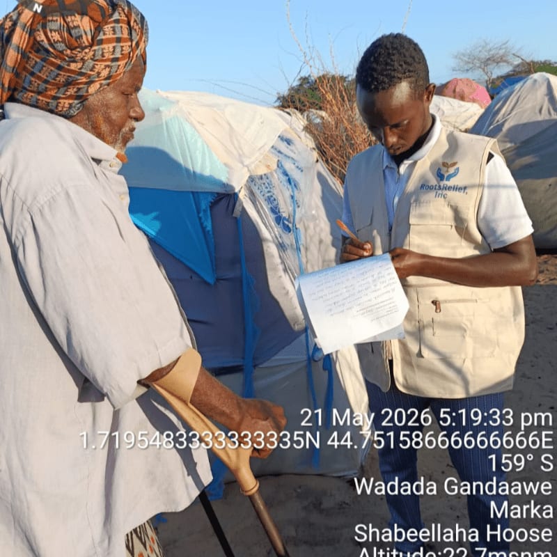 An elderly person with a crutch stands next to a tent while a man in a vest fills out paperwork in the background.