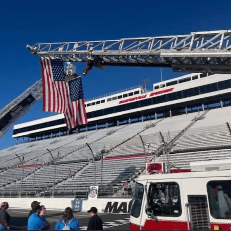 A fire truck and a group of people stand in the foreground before a large, empty grandstand at a speedway, with a fire truck ladder extending upwards displaying two American flags.