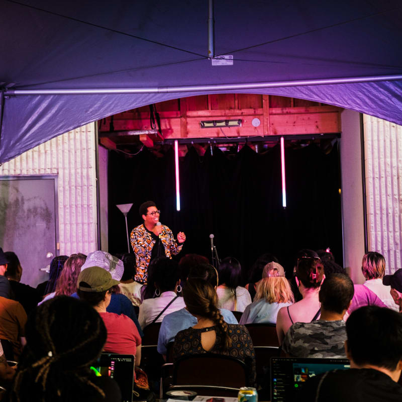 A woman in a colorful jacket speaks into a microphone on a stage under purple lights, while an audience sits in rows facing her.