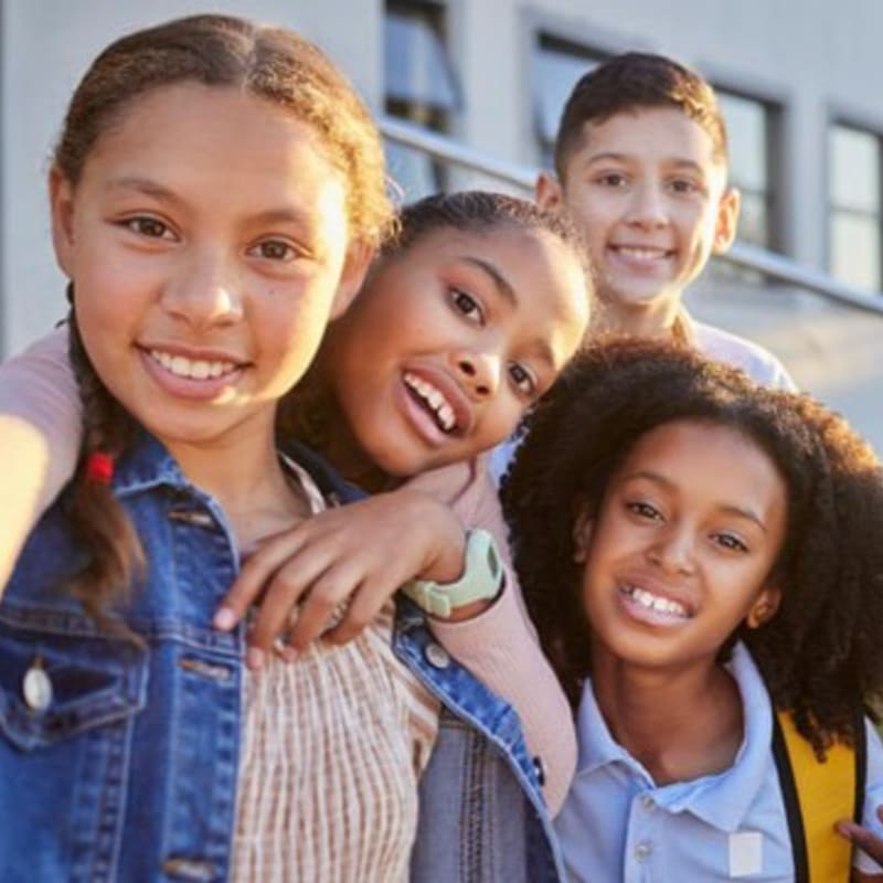 Four smiling children of diverse ethnicities pose for a selfie in front of a blurred building.