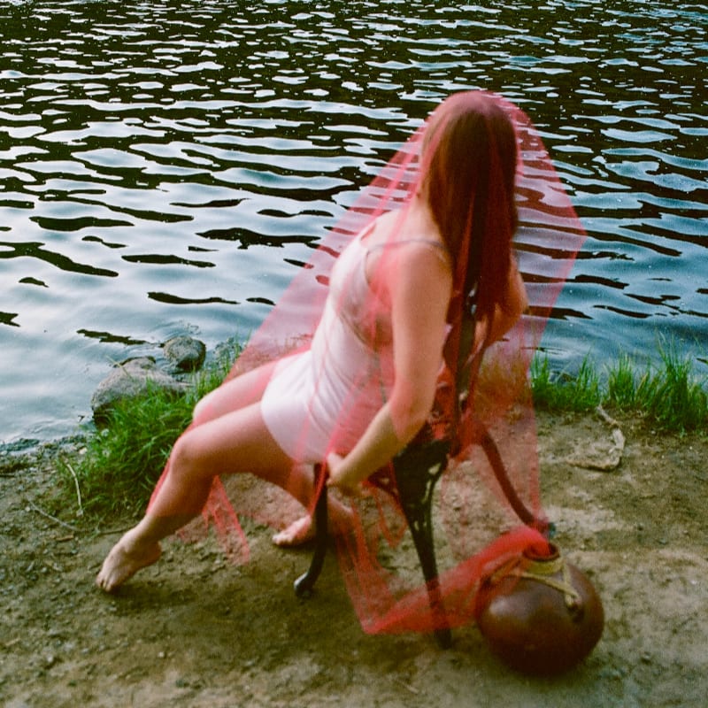 A woman in a light-colored dress and a red veil sits on a chair by the water's edge, with the rippling surface of the lake filling the background.
