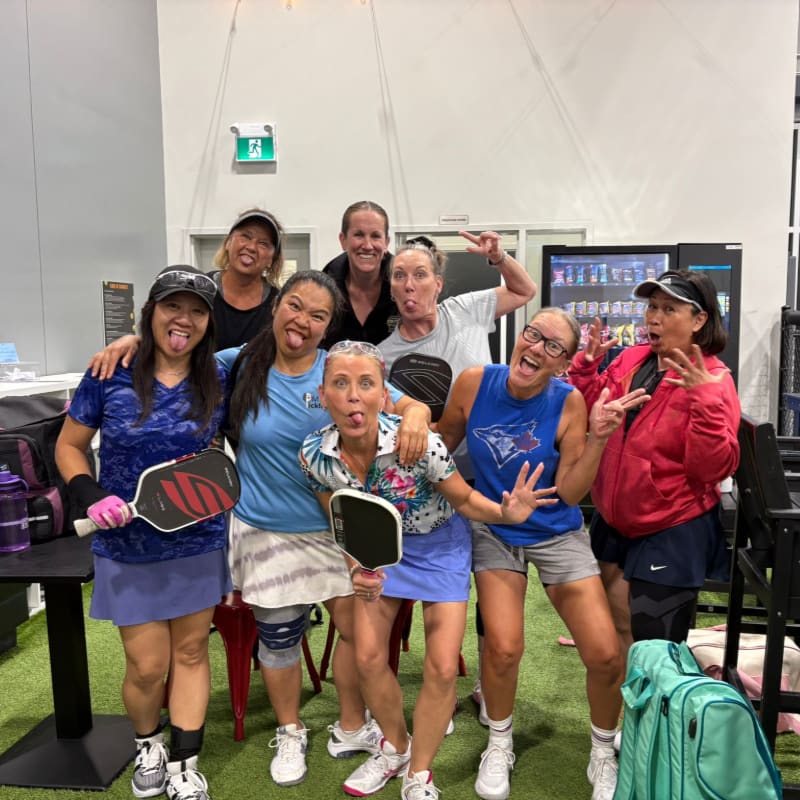 A group of women in athletic wear pose playfully in the foreground, holding pickleball paddles, with a vending machine and gym equipment visible in the background.