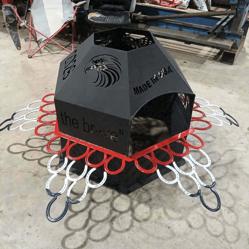 A black metal fire pit with an eagle design and "MADE IN USA" inscribed on it sits on a concrete floor, surrounded by horseshoe wings in red, white, and blue, with a workshop in the background.