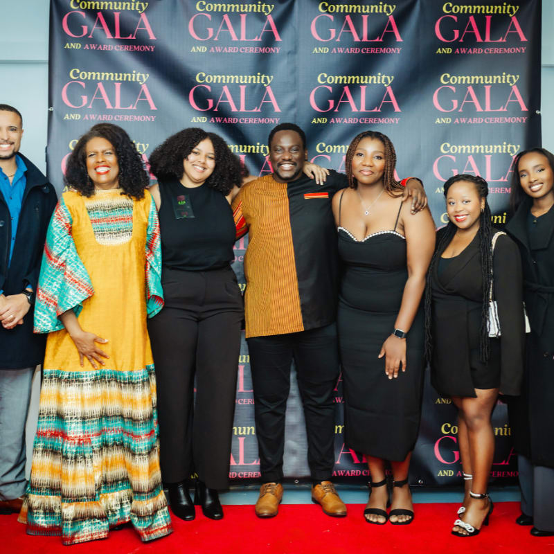 A group of seven people pose on a red carpet in front of a backdrop with "Community Gala and Award Ceremony" repeated.