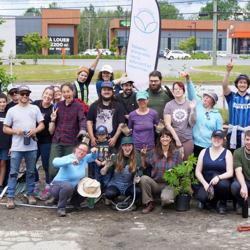 A group of smiling people pose for a photo in front of a building with a green space banner, with some holding gardening tools and plants.