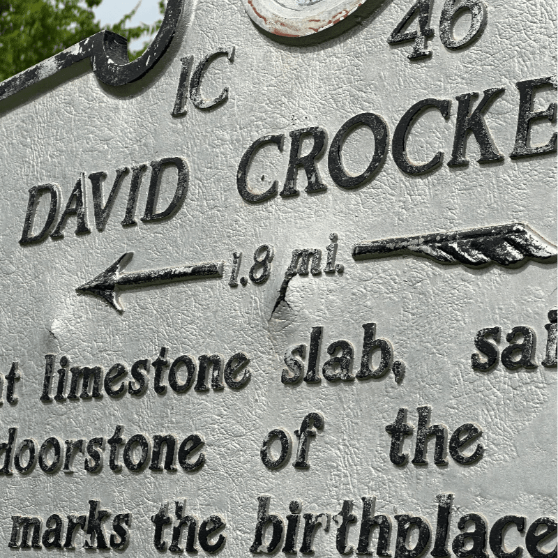 A weathered historical marker in the foreground details David Crockett's birthplace, with a blurred green background suggesting an outdoor setting.