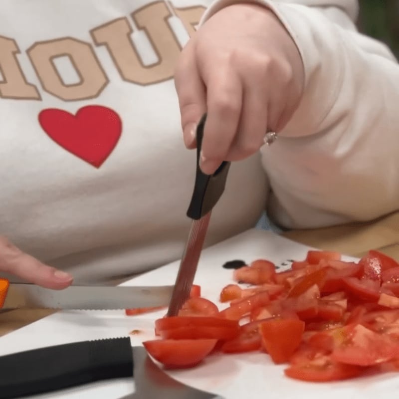 A person in a cream sweatshirt with a red heart is chopping tomatoes on a white cutting board, with a blurred background.