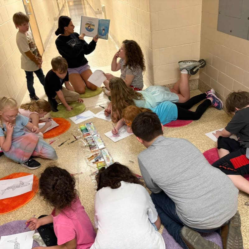 Children sit on colorful mats on a hallway floor, drawing and listening to a teacher read a book in the background.