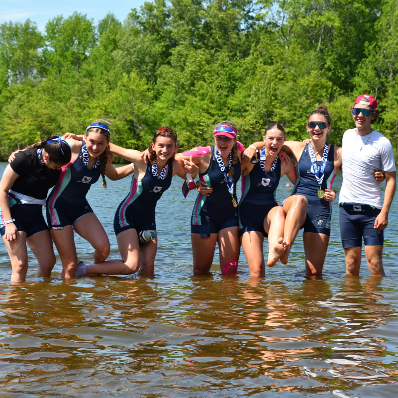 A group of seven young rowers, some wearing medals, stand in shallow water with lush green trees in the background.