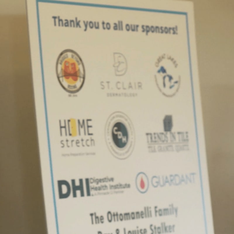 A white sign with blue text and sponsor logos is displayed against a plain, light-colored wall.