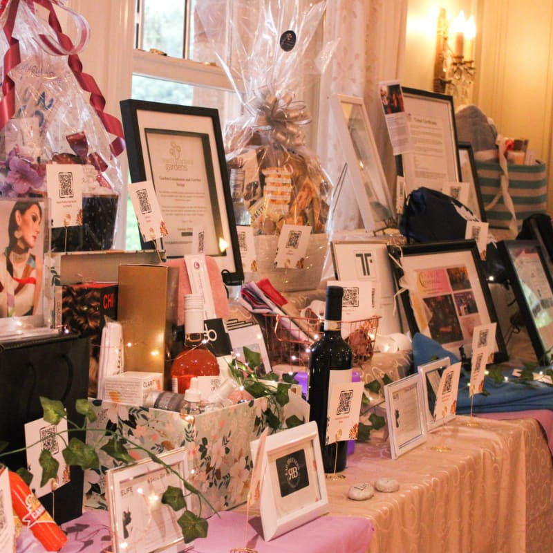 A table laden with gift baskets, framed items, and decorative elements sits in front of a window with sheer curtains.