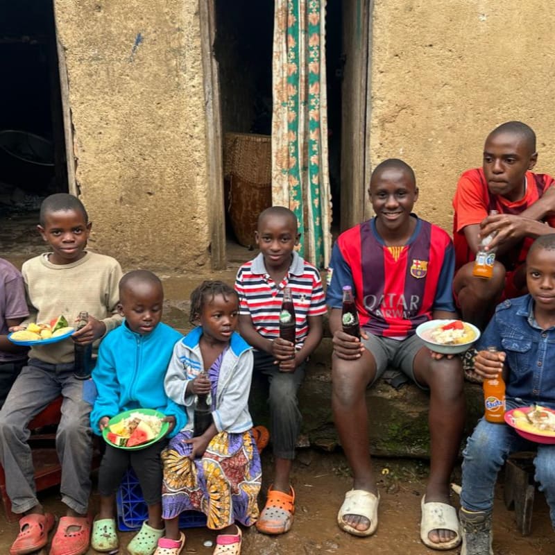 A group of children sit on a step in front of a rustic building, enjoying a meal and drinks.