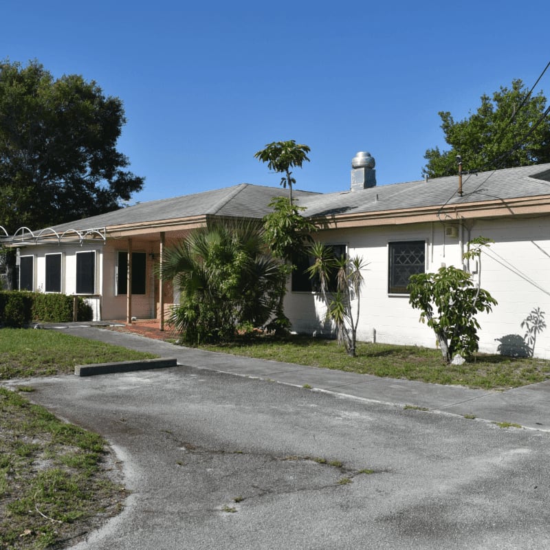 A cracked asphalt driveway leads to a single-story building with lush green trees and bushes surrounding it under a clear blue sky.