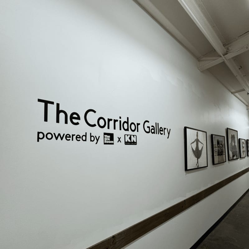 A white wall with "The Corridor Gallery" logo and framed artwork recedes into the distance down a hallway.