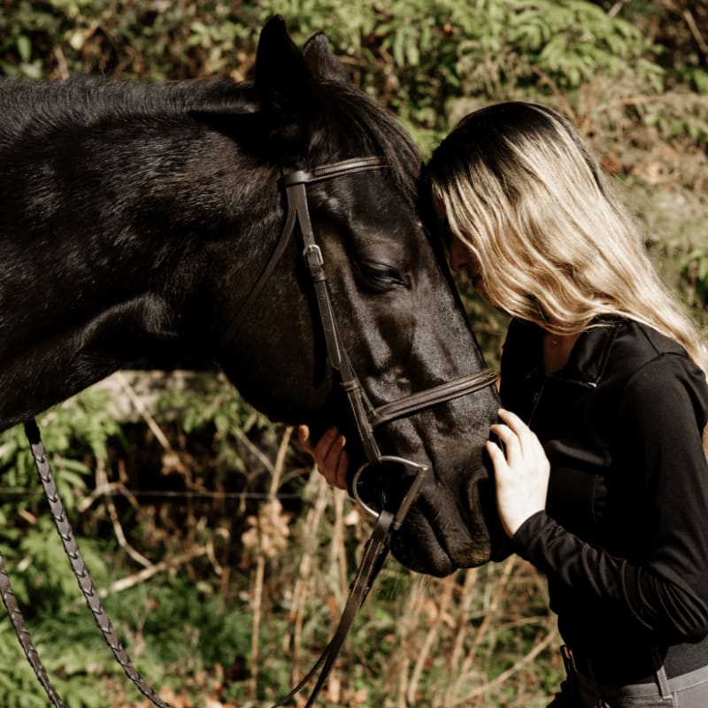 A blonde-haired person gently touches the head of a black horse, with a blurred background of trees and foliage.