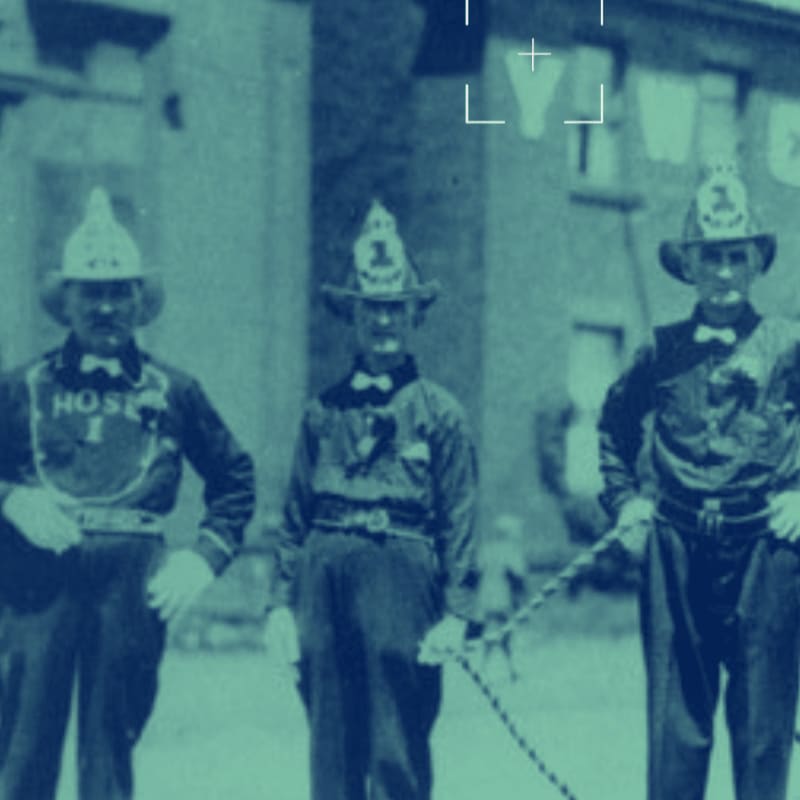 Three men in vintage fire uniforms stand in the foreground, with a blurred building and laundry hanging in the background.