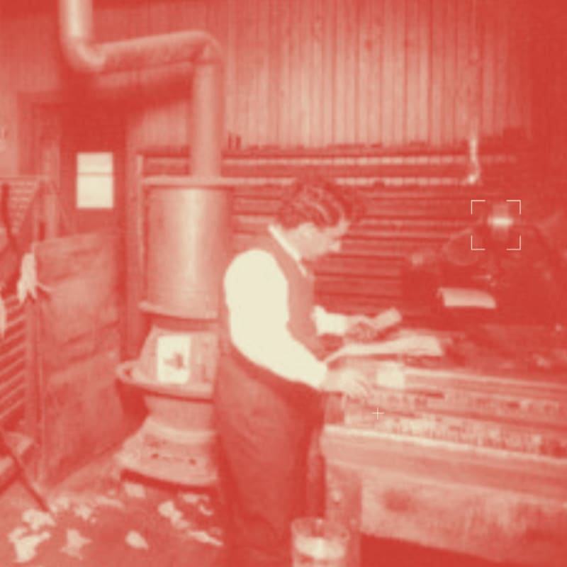 A person in an apron works at a machine in a room with a wood-burning stove and wooden walls.