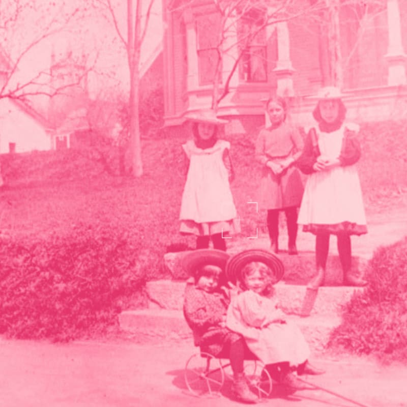 Two children in a wagon in the foreground are posed with four children standing on steps in front of a house in the background.
