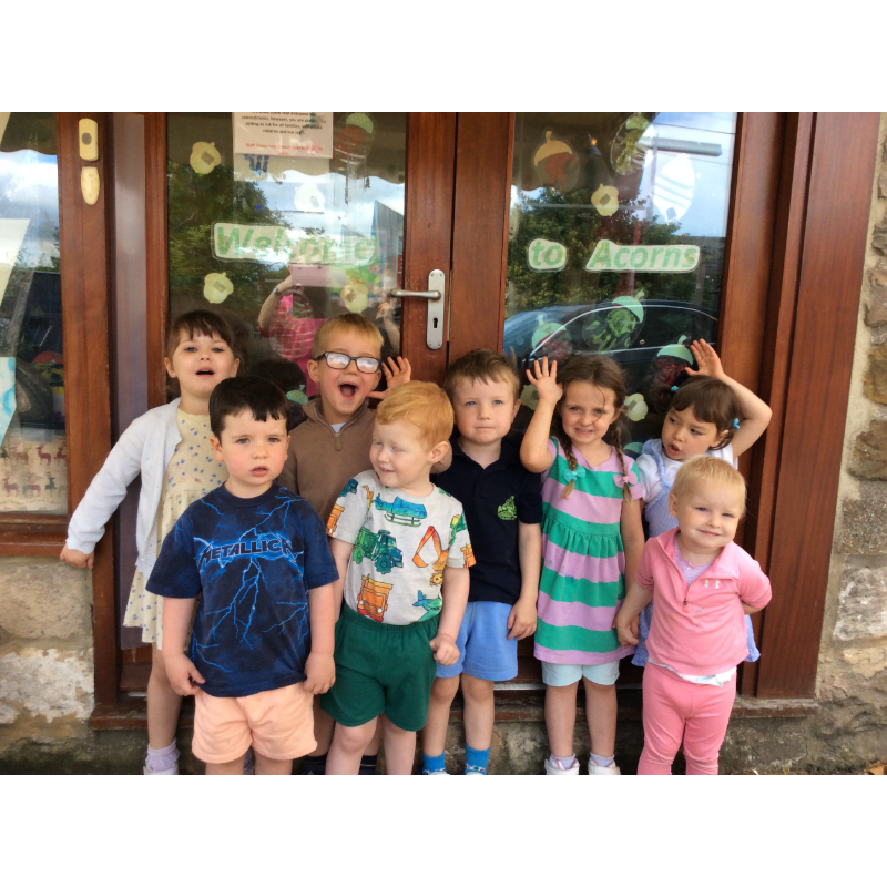 A group of eight young children stand in front of a glass door with "Welcome to Acorns" written on it, with a stone building in the background.