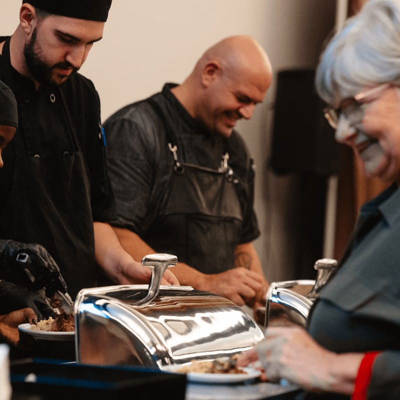 Chefs in black uniforms serve food from chafing dishes to a smiling woman in the foreground, with a blurred background.