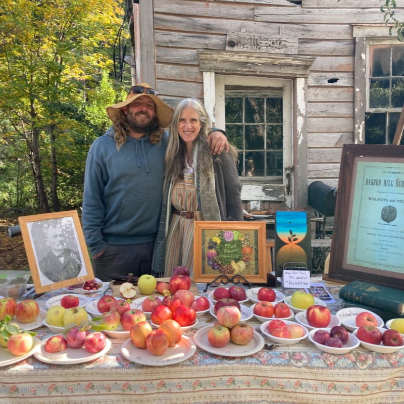 A man and woman stand behind a table laden with apples and framed pictures, with a rustic wooden building in the background.