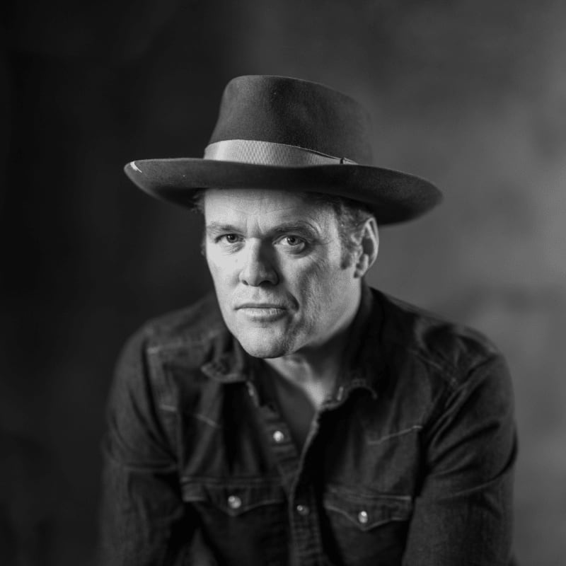 A man in a fedora and denim shirt looks directly at the camera against a dark, textured background.