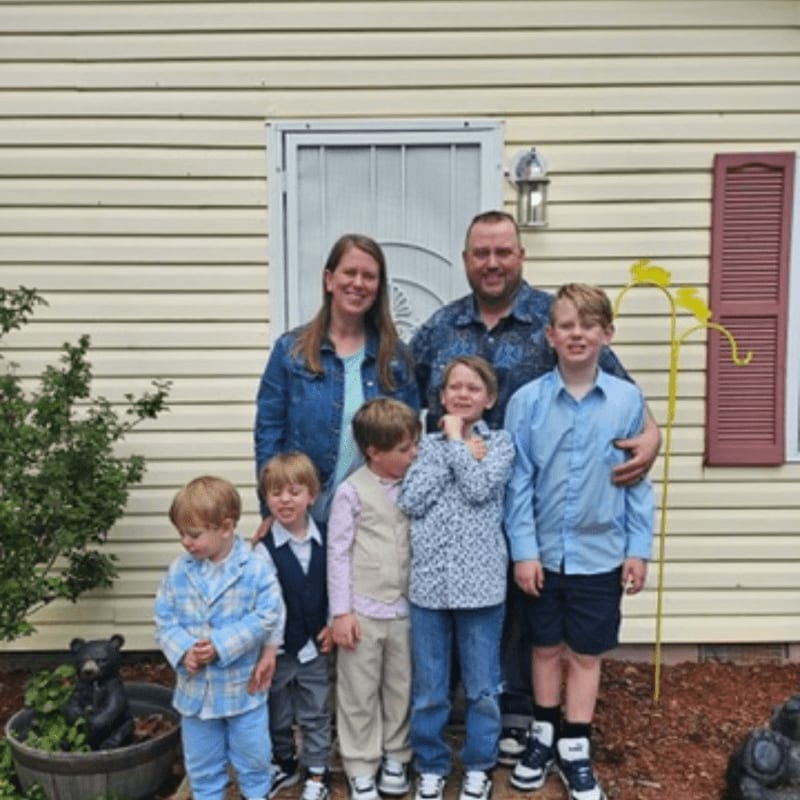 A family of seven poses for a photo in front of a light yellow house with a white door and red shutters.