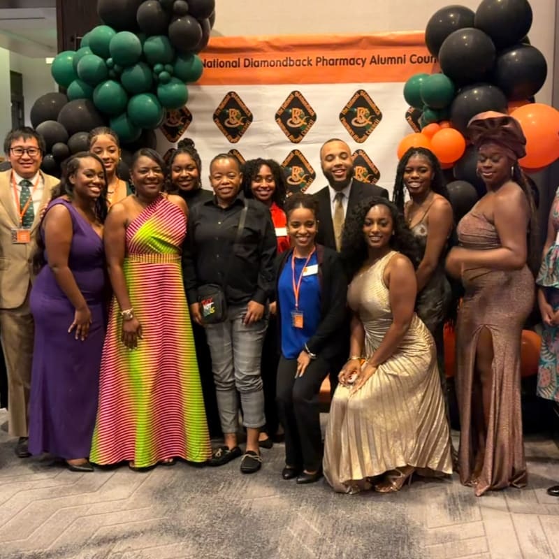 A group of people pose for a photo in front of a banner and balloons at an alumni event.