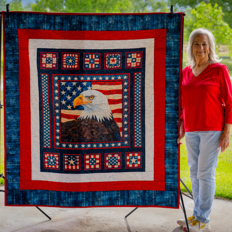 A woman in a red shirt and jeans stands beside a large patriotic quilt featuring an eagle and American flag, with a blurred green background of trees and grass.