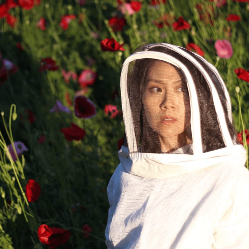 A woman in a beekeeper's suit stands in a field of red poppies.