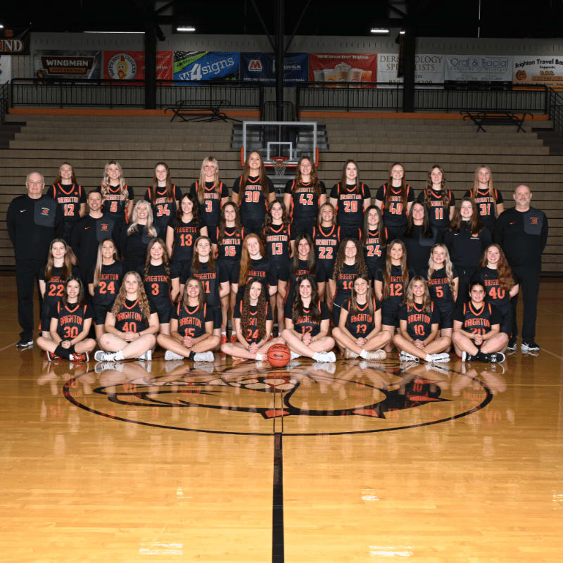 A girls' basketball team poses for a photo on a polished wooden court in front of a gymnasium backdrop.