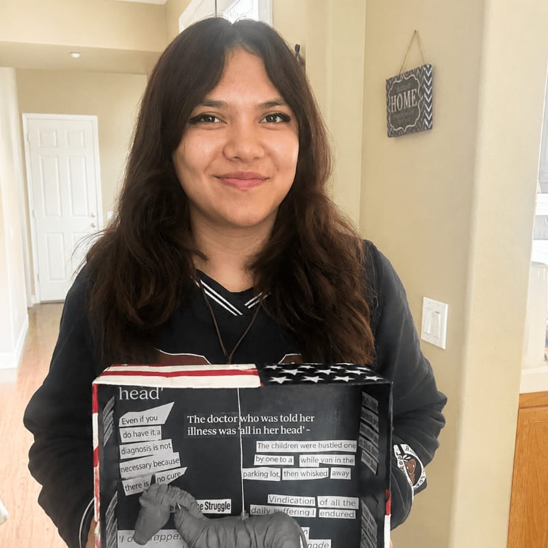 A young woman with long, dark hair smiles as she holds a diorama with text and a zebra sculpture in front of a home interior.