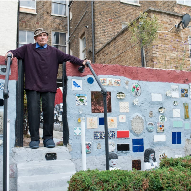 An elderly man stands on a gray staircase in front of a colorful, mosaic-covered wall, with brick buildings in the background.