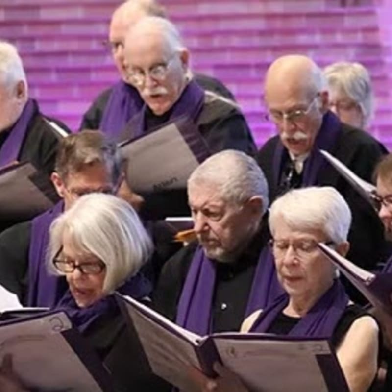 A choir of older adults in purple scarves sings from sheet music in front of a blurred purple background.