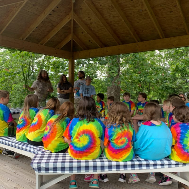 Children in tie-dye shirts sit on a checkered bench under a wooden pavilion, listening to adults in the background amidst lush green trees.